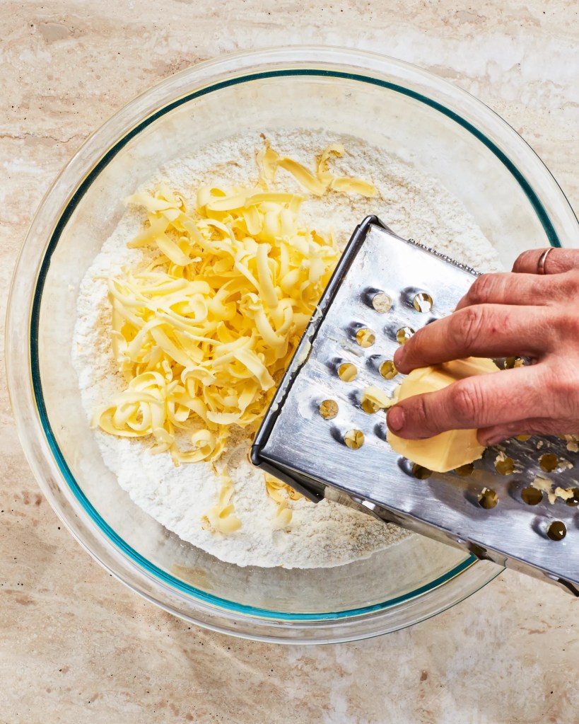 A hand grating frozen butter over a bowl of flour.