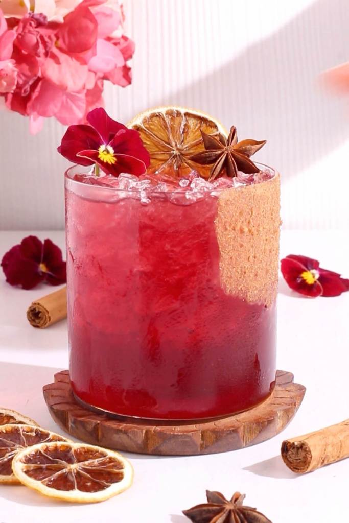 A pin drink in a glass with red pansies, dried lemon, cinnamon stick and anis seed pod.