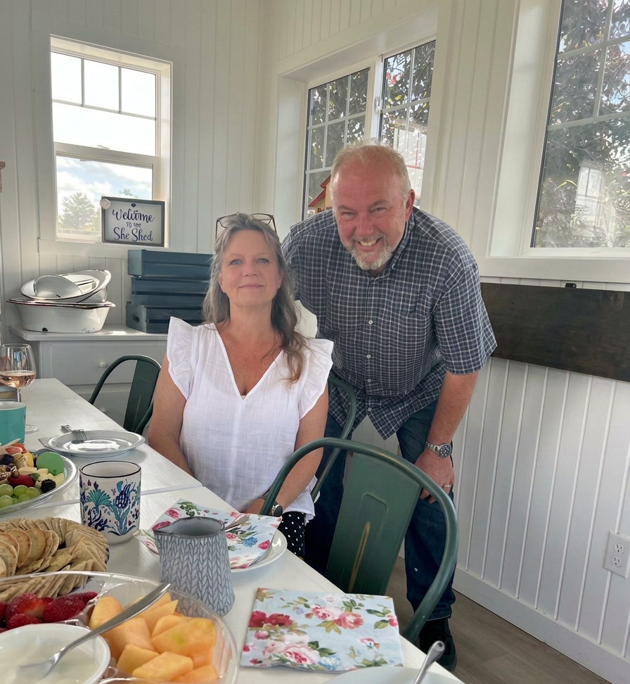 A couple smiling at the camera inside a white Scandi style potting shed. the table is loaded down with food, wine, and coffee. 